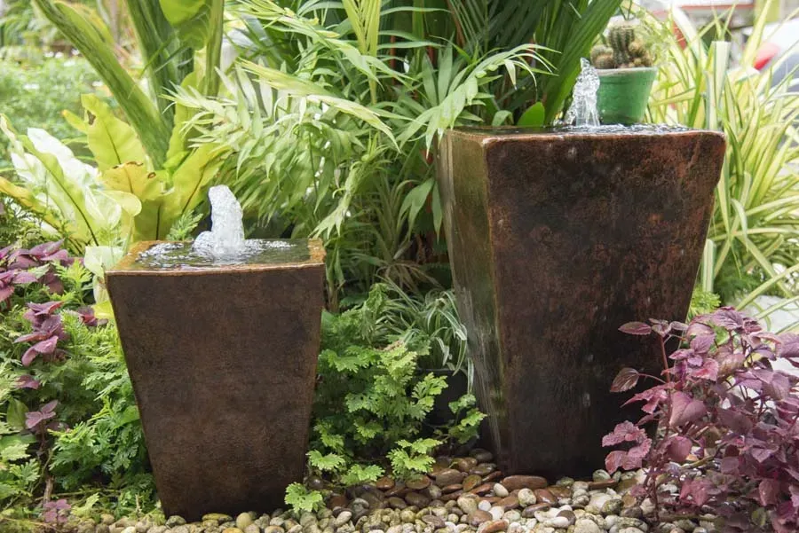 Two bronze-colored, square water fountains in a garden with various green and purple plants and gravel.