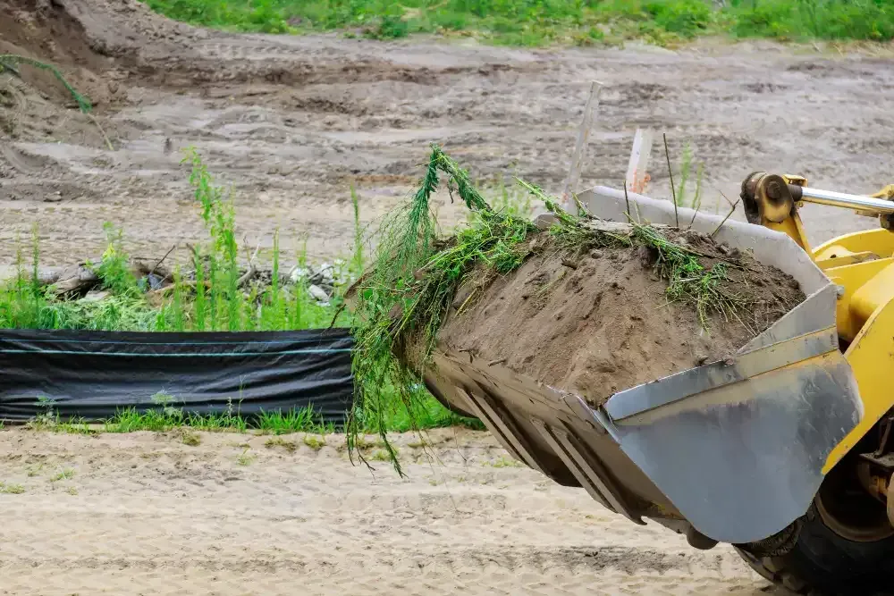 A yellow front-end loader scooping up dirt and weeds at a construction site.