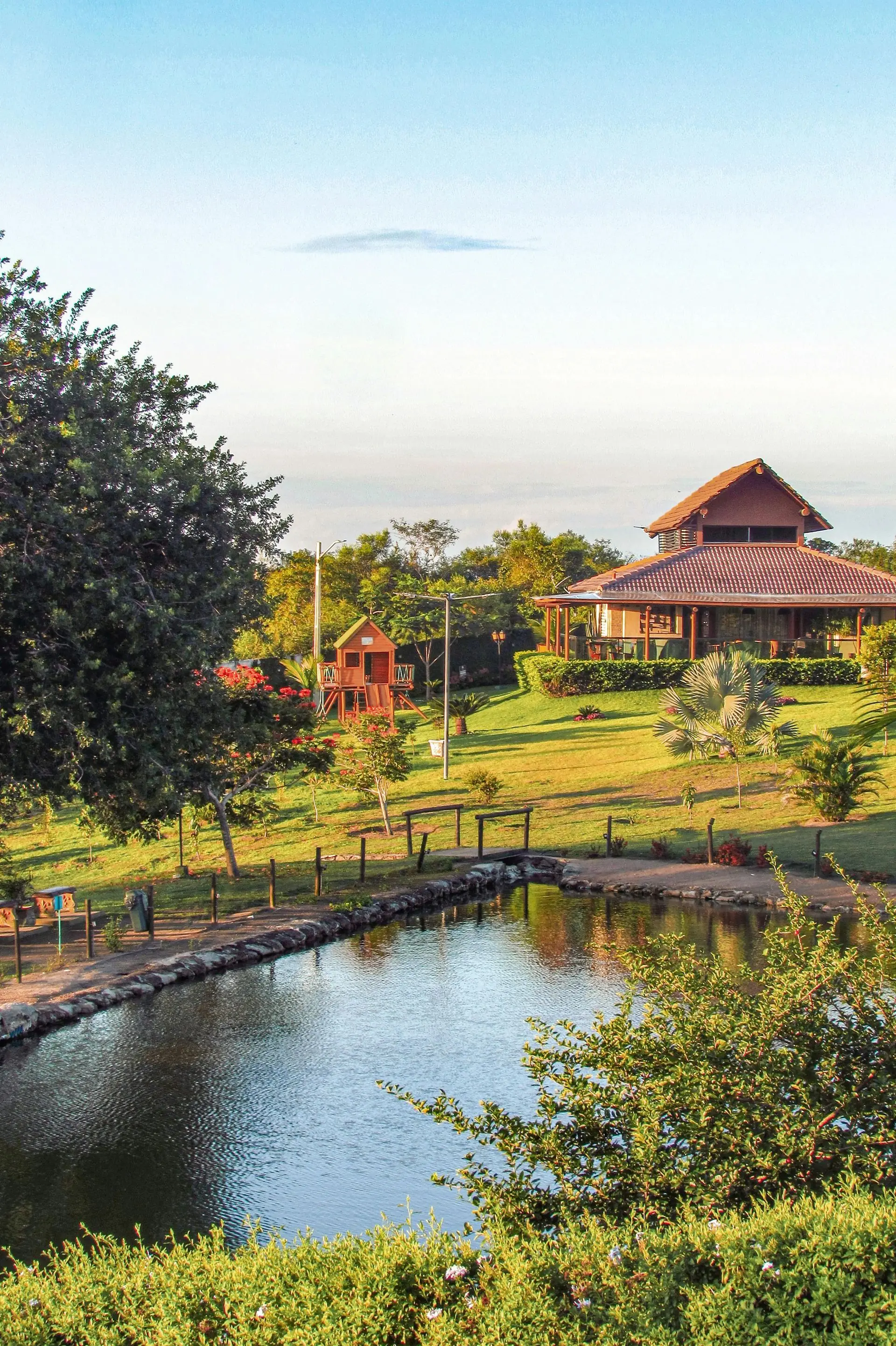 A lake with a wooden bridge in front of a house, set in a sunny, green landscape.