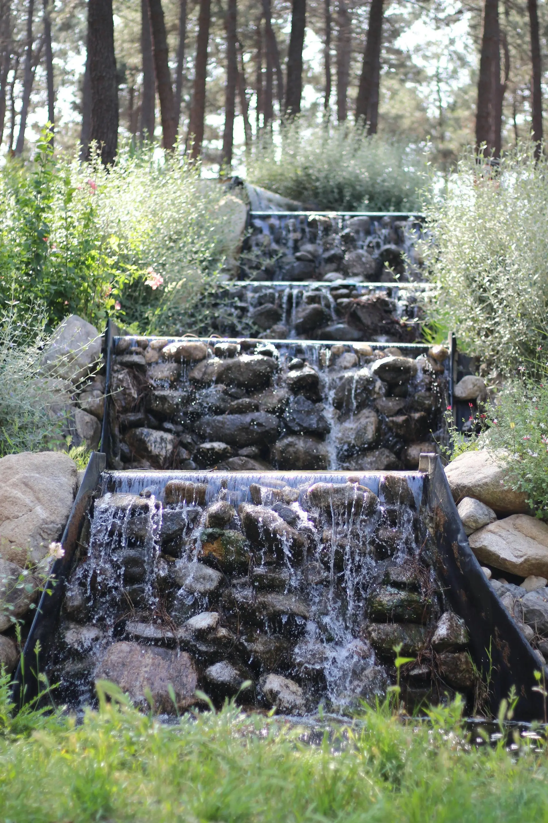 Multi-tiered stone waterfall in a garden, surrounded by greenery and trees. Water cascades down each level.